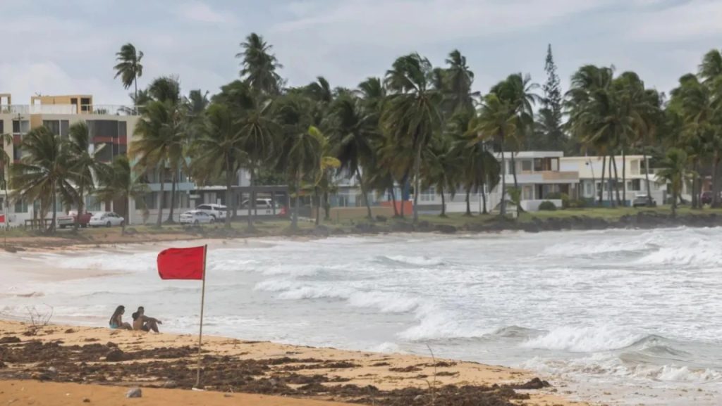 Turistas se sientan en la playa de La Pared mientras la Tormenta Tropical Ernesto agita las olas cerca de Luquillo